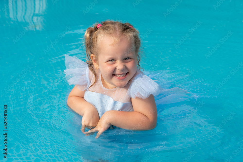 Charming girl 3 years old in a blue dress with blond hair in the pool smiles. Summer holiday concept. Place for text