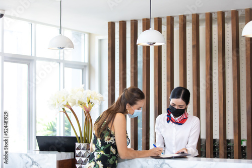 beautiful young asian woman receptionists working at a reception desk and holding key card or passport to customer. People wearing protective face mask prevent covid-19 virus