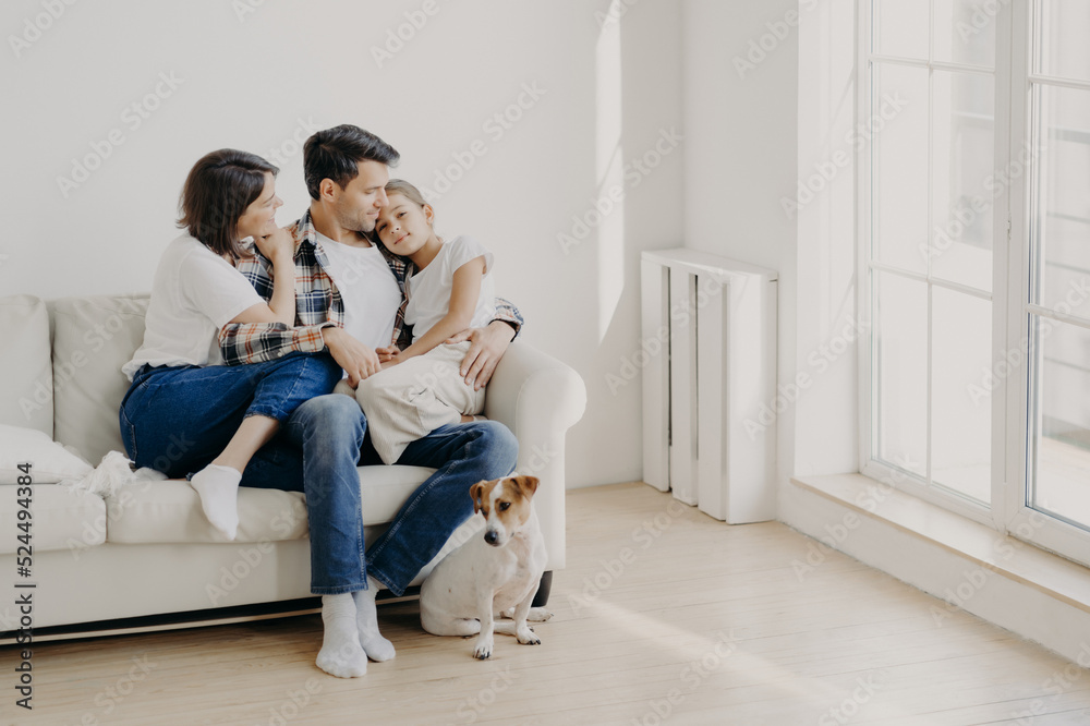 Indoor shot of happy mother and father spend weekend with small daughter, pose all together on comfortable sofa, have fun and pleasant talk with each other. Little dog sits near on floor. Family Indoor shot of happy mother and father spend weekend with small daughter, pose all together on comfortable sofa, have fun and pleasant talk with each other. Little dog sits near on floor. Family