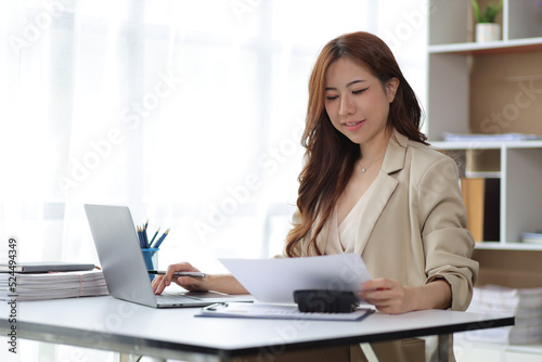 Attractive Asian businesswoman in casual office working on laptop and reading documents.