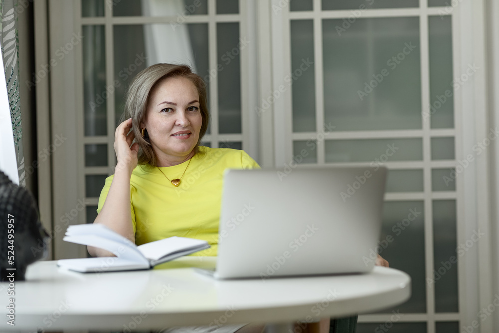 attractive blonde middle-aged woman entrepreneur in a yellow blouse working on a laptop and with documents at home at the table attractive blonde middle-aged woman entrepreneur in a yellow blouse working on a laptop and with documents at home at the table