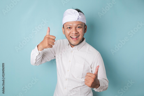 Excited Balinese man wearing udeng or traditional headband and white shirt gives thumbs up hand gesture of approval, isolated by blue background