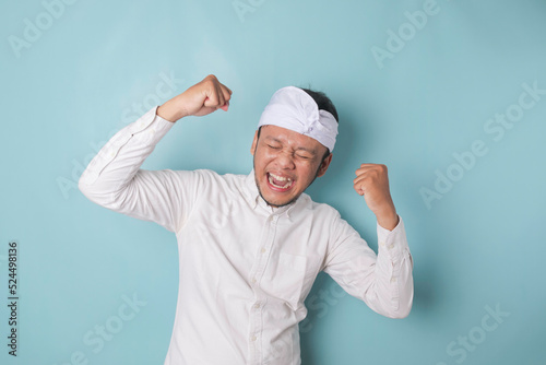 A young Balinese man with a happy successful expression wearing udeng or traditional headband and white shirt isolated by blue background