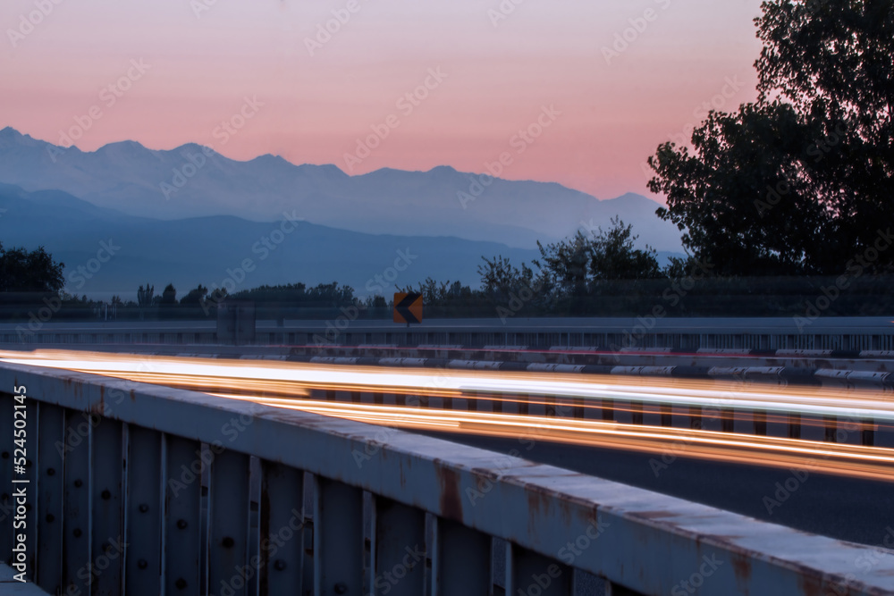 An evening landscape near a curving road with rays of light from passing cars at a long exposure against the background of silhouettes of trees and mountains in blue shades with a sunset pink sky