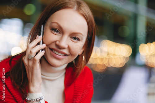 Heashot of lovely European female entrepreneuer has healthy skin, pleasant smile, dressed in red clothes, looks straight at camera, holds modern cell phone, poses outside, blurred background photo