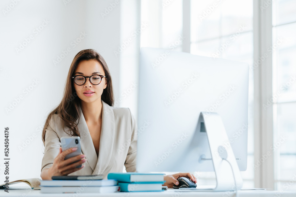 Elegant woman office worker uses mobile phone and computer at one time, sends messages and chats with clients online, monitors news from networks, sits at desktop with pile of notepads and books Elegant woman office worker uses mobile phone and computer at one time, sends messages and chats with clients online, monitors news from networks, sits at desktop with pile of notepads and books
