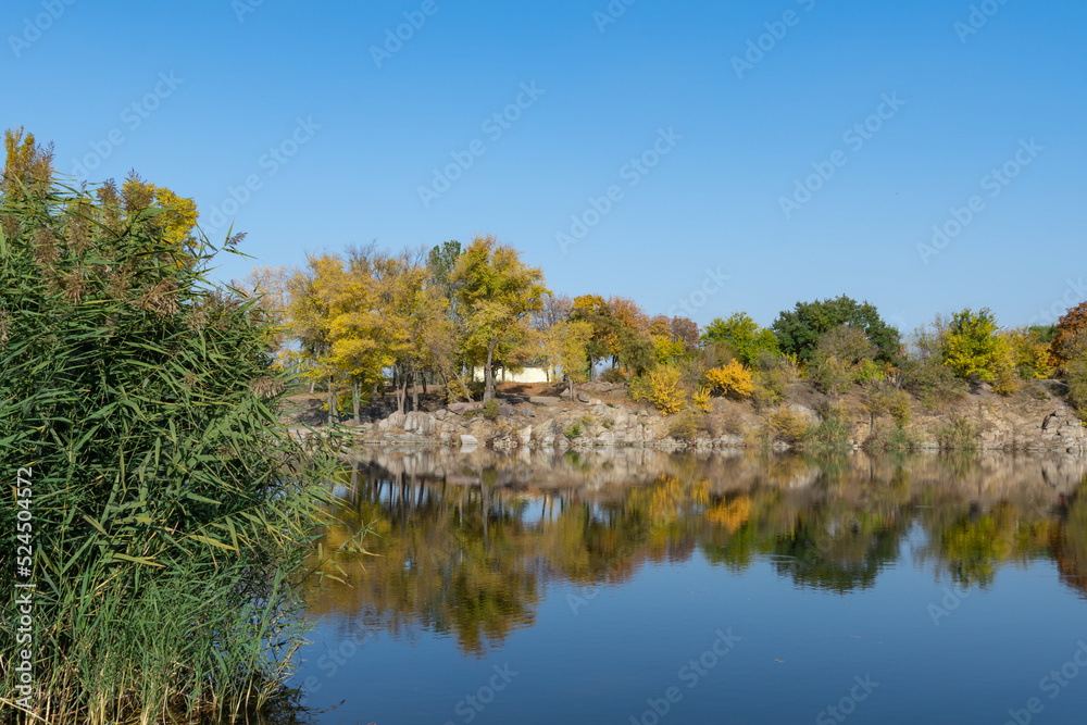 Autumn colors in sunny day on lake in city park. Landscape golden foliage and reflection of trees in calm water. Colorful plants on stone bank of pond and blue sky. Autumn reflections. Fall bright.