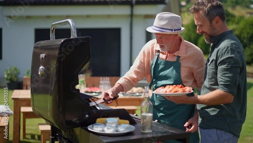 Senior father with adult son grilling outside on backyard in summer family during garden party