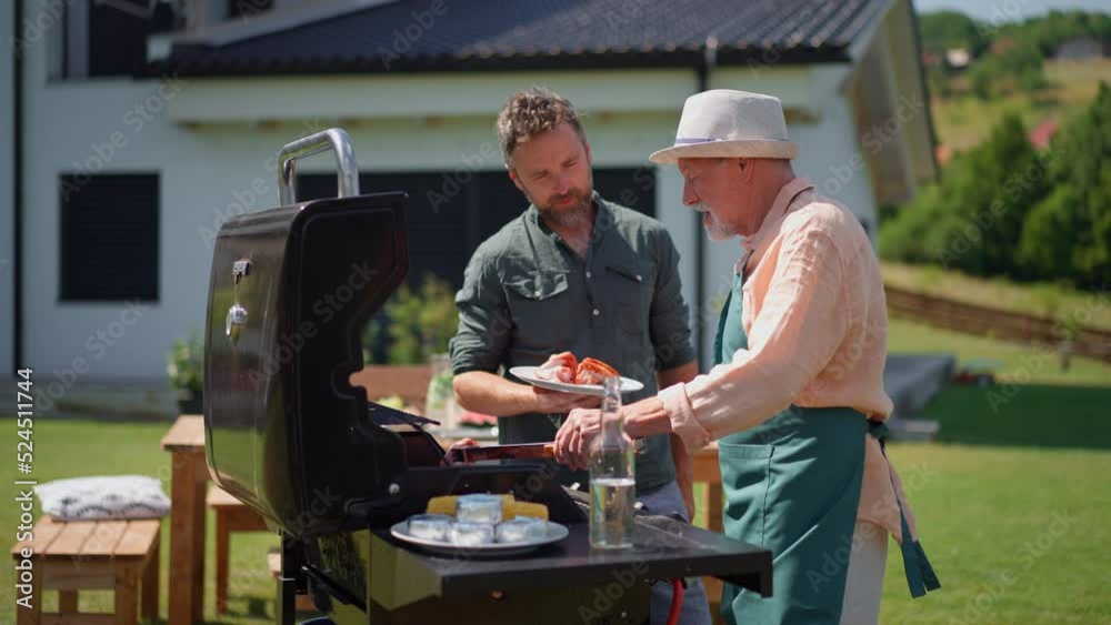 Senior father with adult son grilling outside on backyard in summer family during garden party