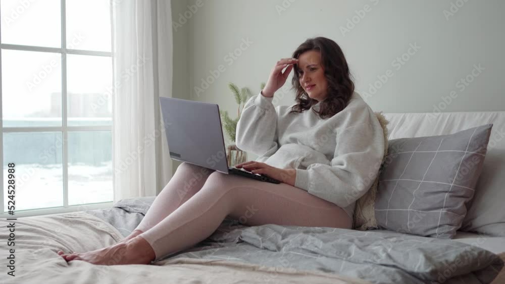 a young pregnant woman in the bedroom sits on the bed and works on a laptop. expecting a baby, motherhood, happiness, pregnancy, a young pregnant woman in the bedroom sits on the bed and works on a laptop. expecting a baby, motherhood, happiness, pregnancy,