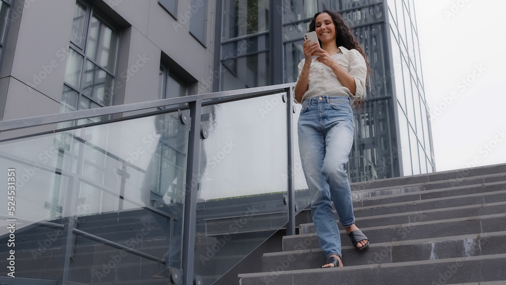 Female 30s slim woman in jeans and shirt standing in city near company building looking phone screen chatting mobile date app. Caucasian businesswoman texting working with smartphone remote outdoors
