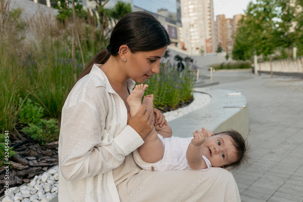Obraz premium Wonderful happy smiling little boy lying on mother's hands in city park. Photo of young mother in white shirt and her kid in city