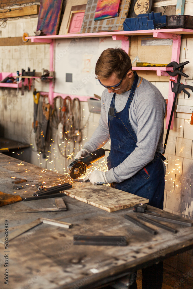 Young metalworker in protective eyewear cutting piece of metal with angle grinder standing at workbench in workshop, side view Young metalworker in protective eyewear cutting piece of metal with angle grinder standing at workbench in workshop, side view