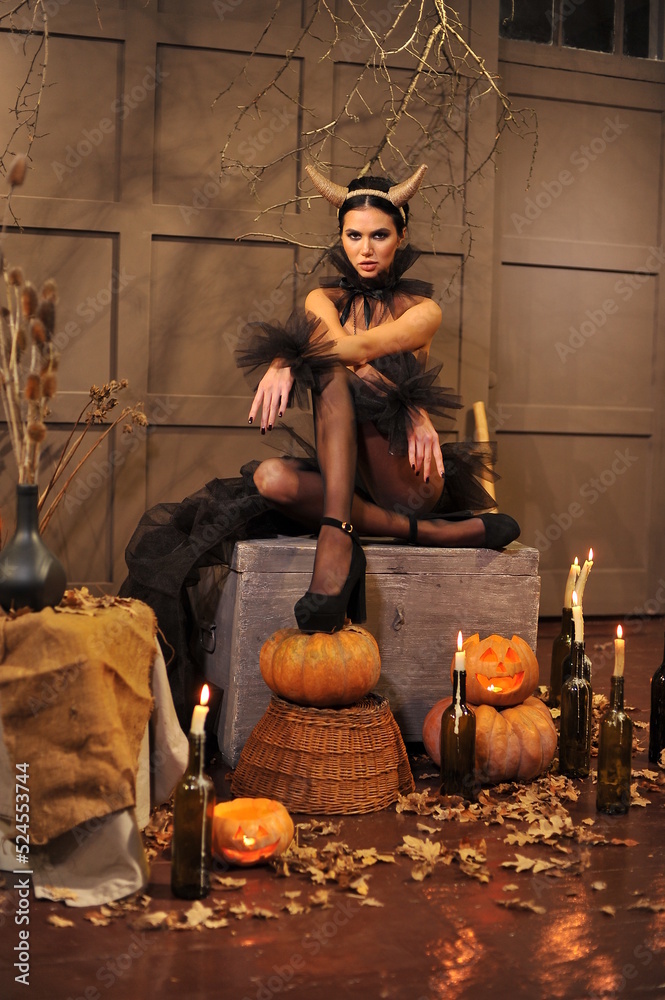 A beautiful brunette with horns and festive Halloween makeup with black manicure and black clothes sits on a chest in her house. Halloween makeup.