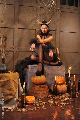 A beautiful brunette with horns and festive Halloween makeup with black manicure and black clothes sits on a chest in her house. Halloween makeup.