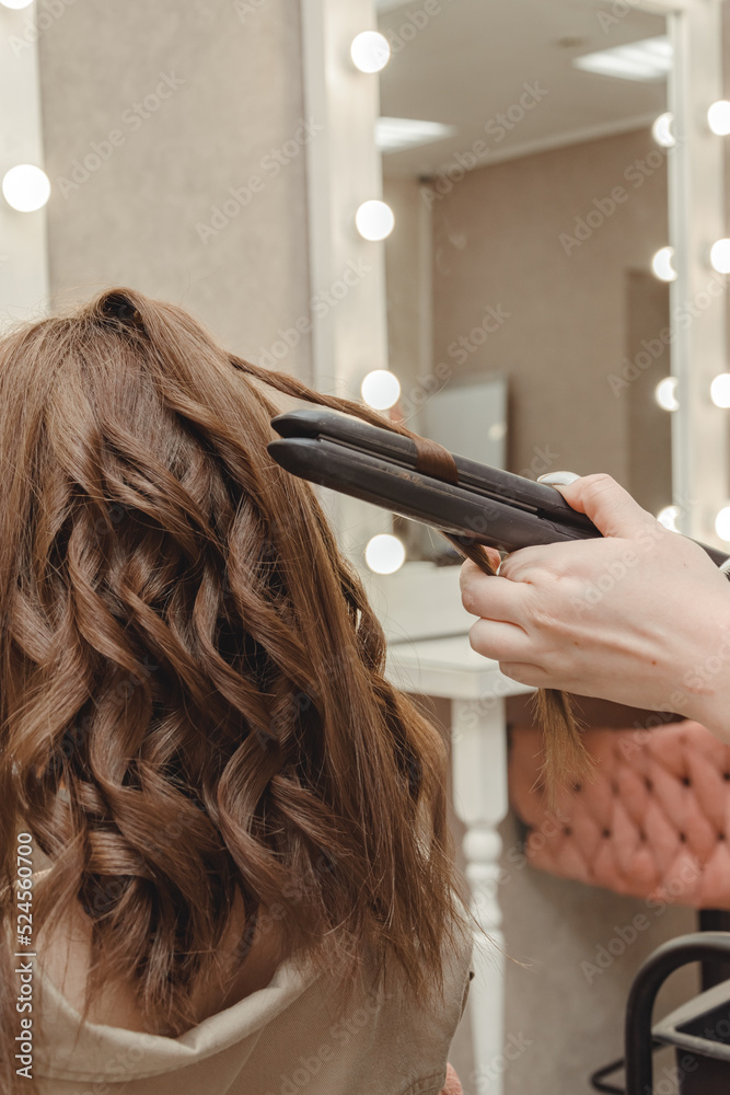 Fototapeta premium girl in beauty salon is doing her hair. women's hair styling on flat iron. blog aesthetics, beauty, hair stylists. hair curling curling iron, space for text.