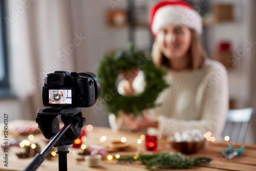 winter holidays, diy and video blogging concept - close up of camera recording happy smiling woman or blogger in santa hat making fir christmas wreath at home