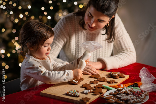 family  cooking and winter holidays concept - happy mother and baby daughter with icing in baking bag decorating gingerbread cookies at home on christmas