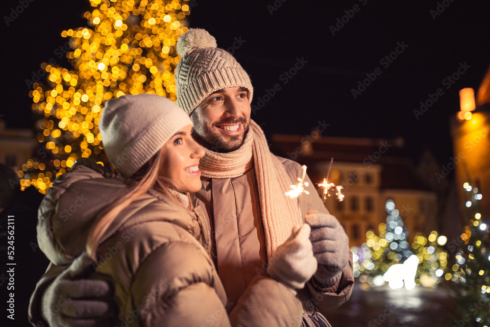 winter holidays and people concept - happy smiling couple with sparklers hugging over christmas lights in city at night