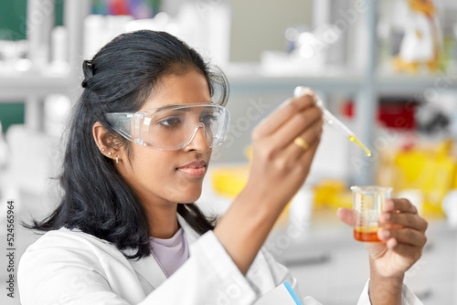 science research, work and people concept - female scientist in goggles with dropper and chemical or test sample in beaker working in laboratory