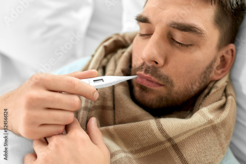 people, health and fever concept - close up of sick man in scarf measuring temperature by thermometer lying in bed