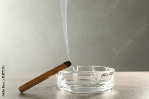 Smoldering cigar near glass ashtray on light grey table, space for text