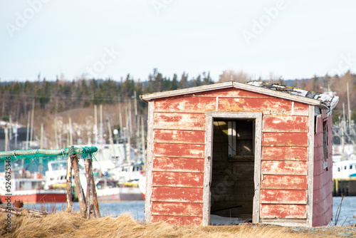 An old red wooden shed with an open door in the foreground and a number of large fishing vessels in the background. The colorful fishing boats are moored at wooden piers. The sky is grey and cloudy. © Dolores  Harvey