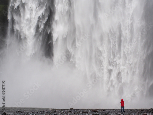 A lone traveler in a red jacket stands before the majestic Skogafoss waterfall in Iceland, showcasing its immense power photo