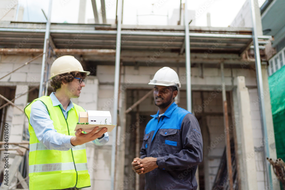 Architect and civil engineer in safety uniform holding house model structure and discuss at construction site on sunny day. Renovate and construction concept. Specialists home project Architect and civil engineer in safety uniform holding house model structure and discuss at construction site on sunny day. Renovate and construction concept. Specialists home project