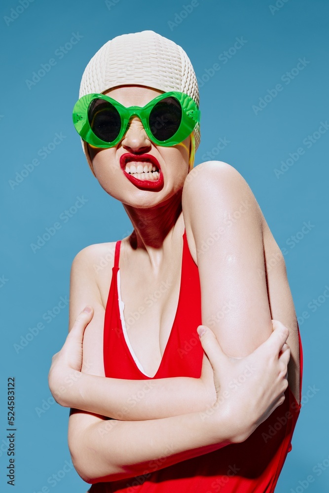 Grimacing unusual girl in a swimming cap and a red swimsuit hugs herself on a blue background in the studio Grimacing unusual girl in a swimming cap and a red swimsuit hugs herself on a blue background in the studio