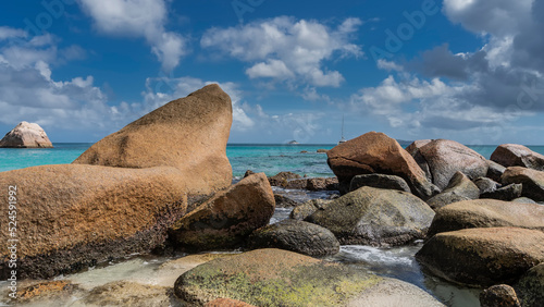 Picturesque granite boulders on a tropical beach. Foam waves between the stones. The yacht is visible in the turquoise ocean. Blue sky with clouds. Seychelles. Praslin. Anse Lazio beach