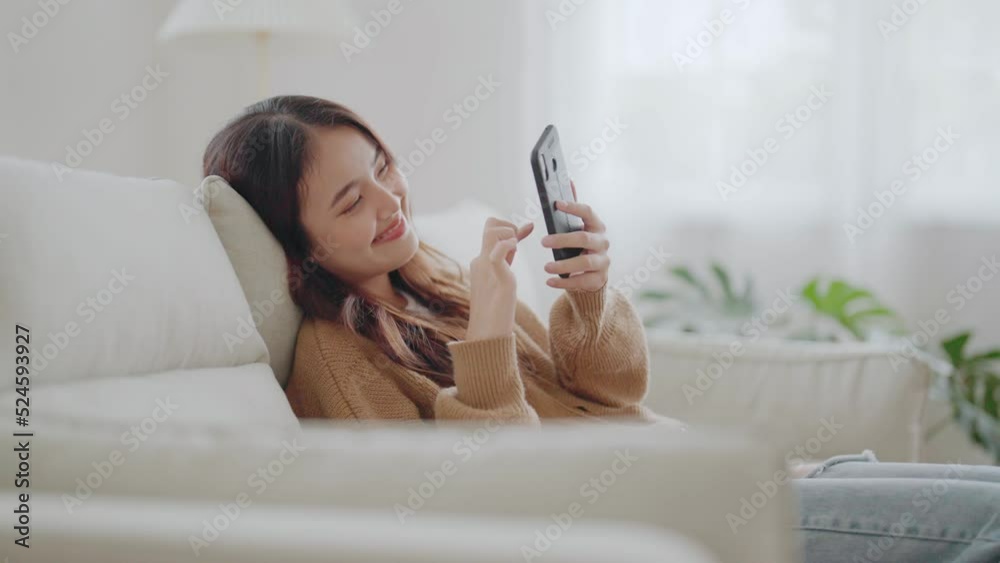 Happy young asian woman relax on comfortable couch at home texting messaging on smartphone, smiling girl use cellphone, browse wireless internet on gadget, shopping online from home Happy young asian woman relax on comfortable couch at home texting messaging on smartphone, smiling girl use cellphone, browse wireless internet on gadget, shopping online from home