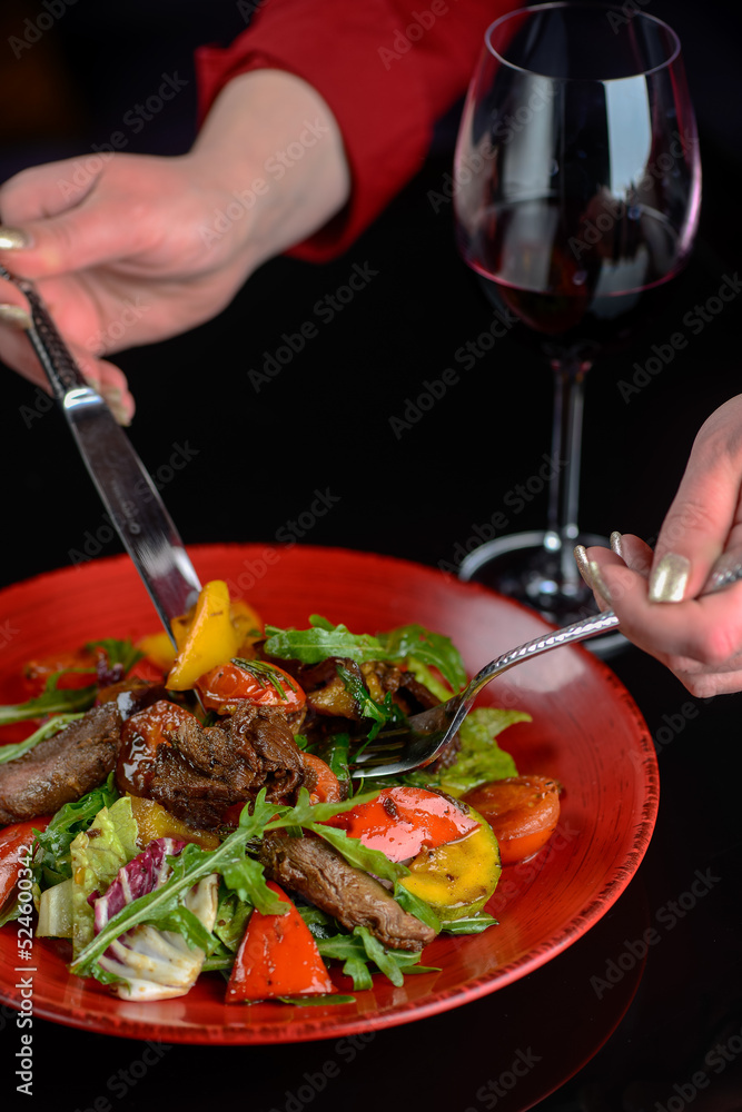 warm salad with veal in a red plate on a dark with utensils in female hands vertical photo

