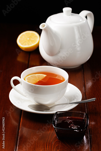 Tea with lemon in a porcelain cup next to a white teapot on a wooden table background. Angle view. Selective focus. Vertical frame orientation.