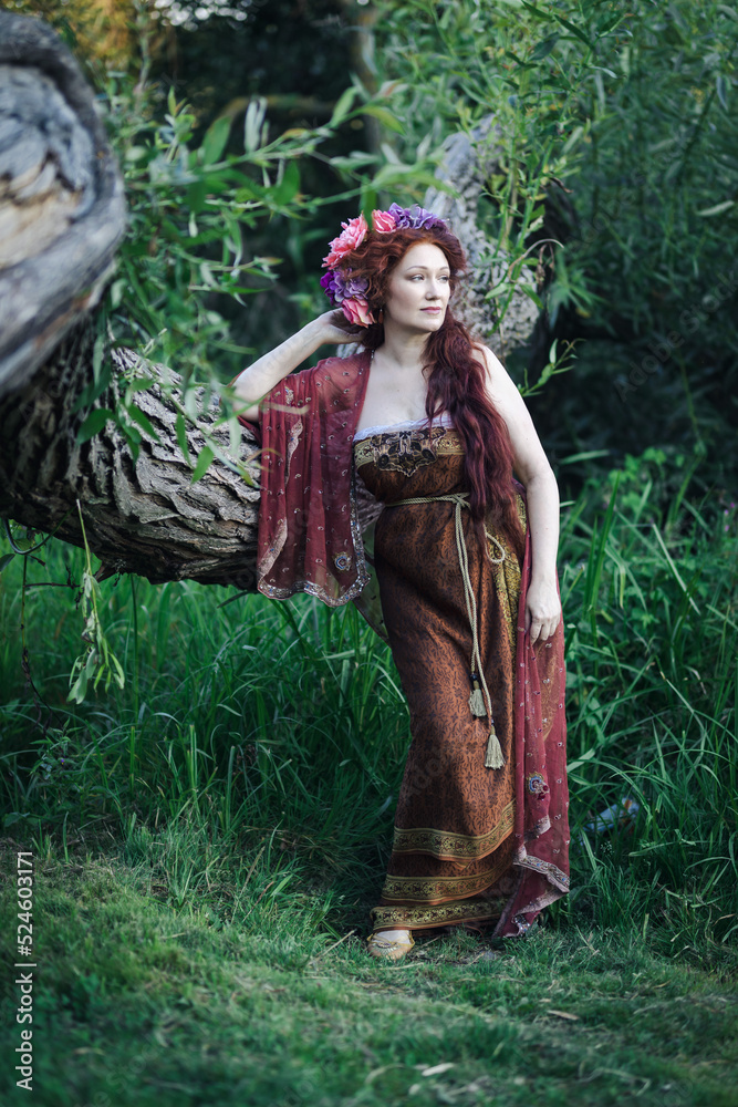 Portrait of a beautiful mature woman dressed in a nymph-style dress inspired by early 20th century artists. A red-haired woman in a wreath near tree. Portrait of a beautiful mature woman dressed in a nymph-style dress inspired by early 20th century artists. A red-haired woman in a wreath near tree.