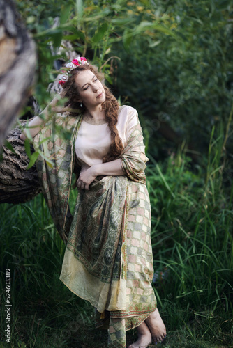 Portrait of a beautiful mature woman dressed in a nymph-style dress inspired by early 20th century artists. Woman posing while leaning on a tree trunk.