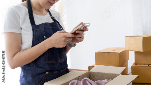Hands of Asian woman entrepreneur holding smartphone with cardboard boxes, computer laptop, online selling equipments. Packing, business and technology concept. Top view, copy space