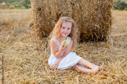 a little curly-haired girl is sitting by a haystack in a field with a green apple in her hands
