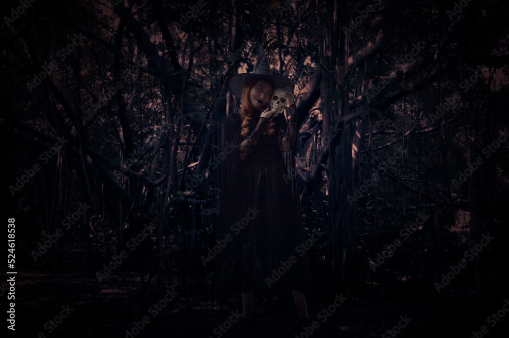 Halloween witch holding a skull standing over spooky dark forest with tree, leaves and vine, Halloween mystery concept