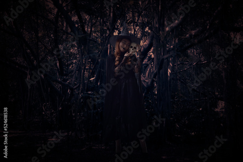 Halloween witch holding a skull standing over spooky dark forest with tree, leaves and vine, Halloween mystery concept