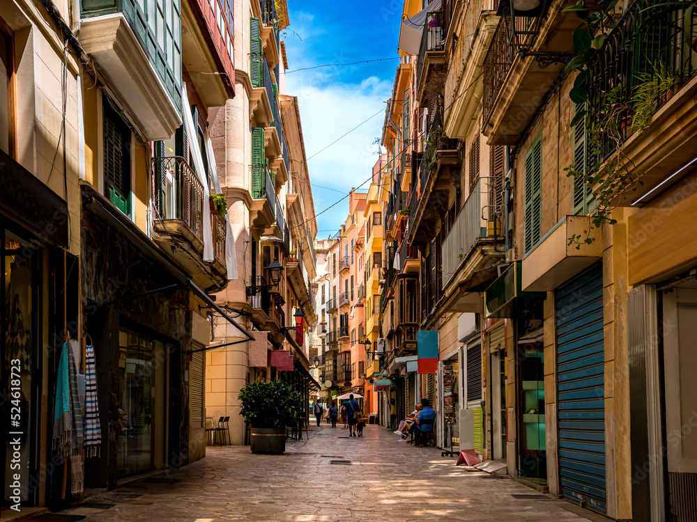 Street view of empty Carrer de l'Argenteria in a mediterranean shopping district of Palma de Mallorca with unrecognizable pedestrians, small businesses, coffee shops and restaurants on a summer day.