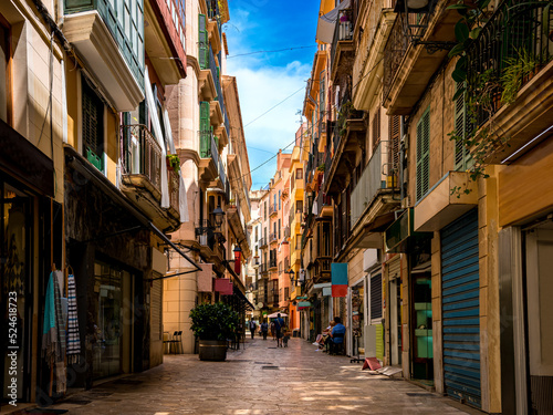 Fototapeta Naklejka Na Ścianę i Meble -  Street view of empty Carrer de l'Argenteria in a mediterranean shopping district of Palma de Mallorca with unrecognizable pedestrians, small businesses, coffee shops and restaurants on a summer day.