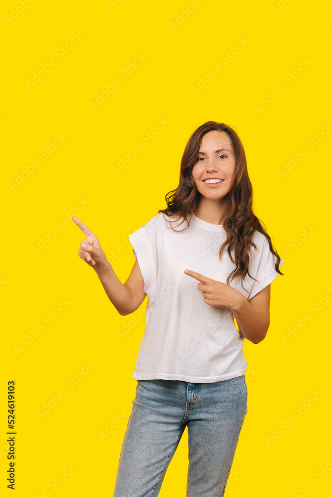 Woman with wavy brown hair is smiling at the camera and pointing at a free space, wears a white shirt while standing over yellow background