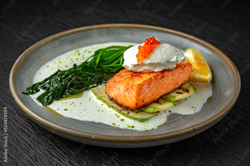 Grilled salmon fillet with zucchini, spinach, cream cheese, garnished with red caviar with cream sauce in a ceramic plate on a dark textured background. Restaurant menu Isolated on black Grilled salmon fillet with zucchini, spinach, cream cheese, garnished with red caviar with cream sauce in a ceramic plate on a dark textured background. Restaurant menu Isolated on black
