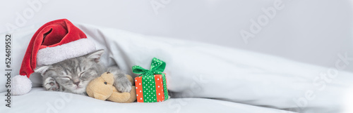 Photo Funny kitten wearing red santa's hat sleeps with gift box under white blanket and hugs favorite toy bear