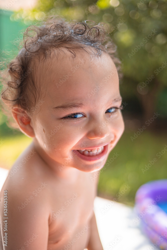 Happy kid playing in an inflatable pool in the backyard. Curly toddler smiling and playing with water Happy kid playing in an inflatable pool in the backyard. Curly toddler smiling and playing with water