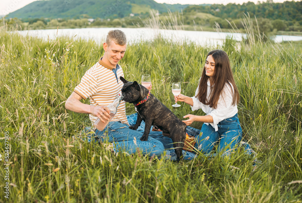 young happy couple having picnic with their french bulldog outdoors in the countryside during beautiful sunset. Romantic date near the lake young happy couple having picnic with their french bulldog outdoors in the countryside during beautiful sunset. Romantic date near the lake