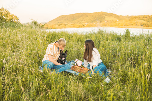 young happy couple having picnic with their french bulldog outdoors in the countryside during beautiful sunset. Romantic date near the lake