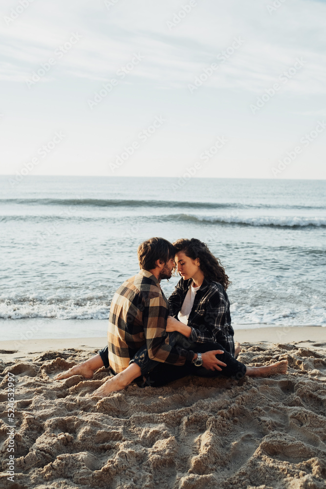 Young woman and man hugging while sitting together on the seashore with sea waves on the background at sunrise
