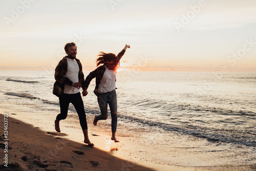 Happy young couple holding by hands and running along the coastline, woman and man meeting sunrise on the sea together